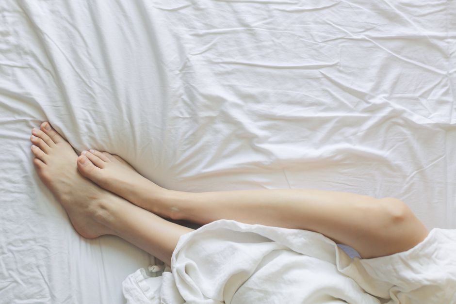 Illustration of a person lying on their back with legs elevated up a wall in Viparita Karani, arms relaxed at sides, in a soft bedroom with natural daylight from a window